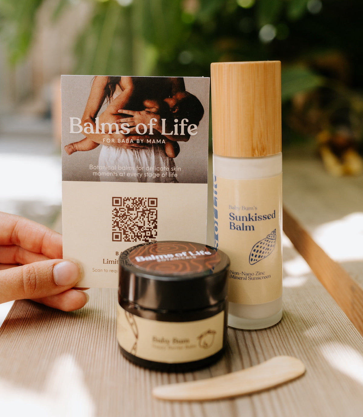 Hand holding a Balms of Life card behind a jar of Baby Bum nappy barrier balm with a wooden spatula